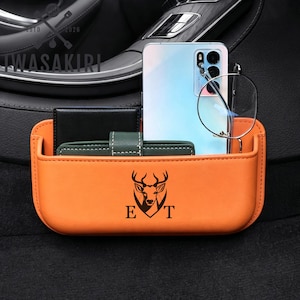 Peut inclure: Un organisateur d'espace de siège de voiture en cuir orange avec un logo de tête de cerf imprimé en noir et les lettres E et T. L'organisateur contient un smartphone bleu clair, une paire de lunettes rondes, un portefeuille noir et un petit étui vert.