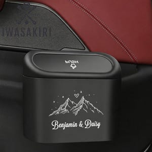 以下が含まれることがあります： プッシュ式開閉蓋付きのマットブラックな車用ゴミ箱。前面には山、星、ハートの白い線画が描かれ、その下に「Benjamin & Daisy」という文字が白い筆記体でプリントされています。コンパクトなデザインで、車内のドアポケットなどに設置可能です。