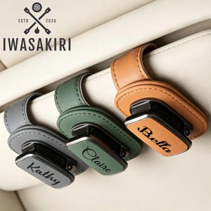 May include: Three car visor clips in gray, green, and tan, each featuring a leather-like strap and a rectangular metal plate engraved with the names Kathy, Claire, and Bella. The logo IWASAKIRI with a crossed wrench icon is visible at the top.