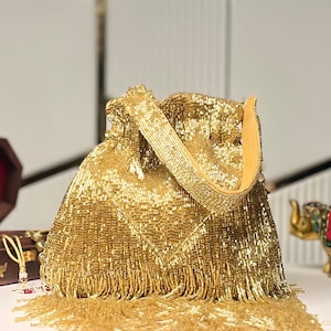 May include: A gold beaded handbag featuring a structured body covered in shimmering metallic beads, a wide shoulder strap, and a long fringe trim along the bottom edge that pools onto the surface. The bag has a drawstring-style closure.