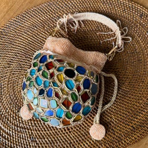 May include: A drawstring pouch bag featuring a surface covered in multi-colored, translucent, faceted glass-like stones in shades of blue, green, red, and yellow. The bag has a pale peach beaded handle and matching drawstring cords with beaded ends.