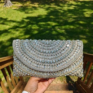May include: A rectangular clutch bag covered in clear, faceted crystals arranged in concentric semicircular patterns. The bag features a flap closure with a dense, shimmering surface of reflective stones that catch the light, creating a brilliant, textured appearance across the entire front exterior.