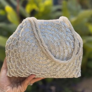 Puede incluir: Un bolso estructurado cubierto con intrincados abalorios plateados y dorados dispuestos en un patrón repetitivo de escamas en forma de abanico. El bolso cuenta con un asa de cuentas a juego y un marco rectangular, con una textura creada por densas cuentas metálicas brillantes.
