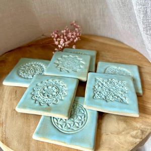 Könnte beinhalten: Eine Sammlung quadratischer Keramikfliesen mit hellblauer Glasur, die jeweils ein einzigartiges geprägtes geometrisches oder florales Muster aufweisen. Die Fliesen haben eine glatte, matte Oberfläche mit sichtbarer Textur und sind überlappend gestapelt.
