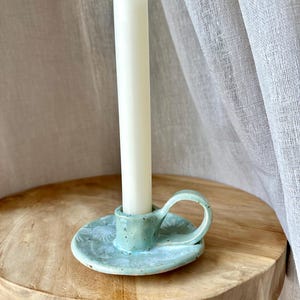 Può includere: Un portacandele in ceramica azzurro con finitura maculata e una piccola impugnatura, caratterizzato da un motivo floreale bianco sulla base. Una singola candela affusolata bianca, alta e sottile, è inserita al centro del portacandele.