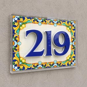 May include: A rectangular house number sign displaying the digits 219 in raised, deep blue lettering. The background features a cream-colored center framed by a decorative border of yellow lemons, blue flowers, and green foliage on a white tile base.