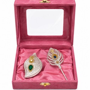 May include: A pink velvet jewelry box with a clear lid containing two silver-toned items: a leaf-shaped container with a green teardrop stone and a pearl, and a peacock feather-shaped accessory featuring a blue stone, a pearl, and gold accents.