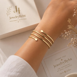 May include: A gold-toned spiral wrap bracelet featuring three thin, polished bands that coil around the wrist, each ending in a small, rounded teardrop-shaped terminal. The bracelet is presented alongside branded packaging with the text JewelryByIst.