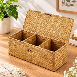 May include: A rectangular woven storage box with a hinged lid and three internal compartments. The box is made of light brown natural fibers in a tight basket weave pattern and features a small dark brown wooden toggle closure on the front.