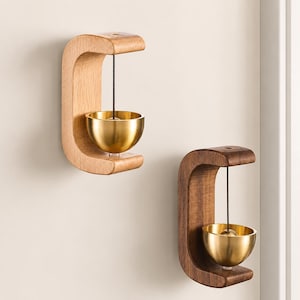 May include: Two wall-mounted doorbells featuring a C-shaped wooden frame. One frame is light-toned wood and the other is dark-toned wood. Each doorbell has a polished gold-colored metal bowl suspended by a thin black cord from the top of the frame.