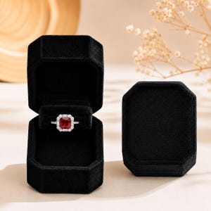 May include: A silver ring featuring a large, square-cut red gemstone surrounded by a halo of small, clear white stones, displayed inside an open, black velvet, octagonal jewelry box next to a matching closed black velvet octagonal box.