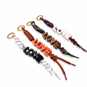 Peut inclure: Quatre porte-clés avec des cordons tressés en blanc, orange, marron et violet. Chaque porte-clé possède un anneau en métal doré et quatre lettres en bloc formant le mot LOVE en noir, orange, marron et blanc, avec des perles et des nœuds.
