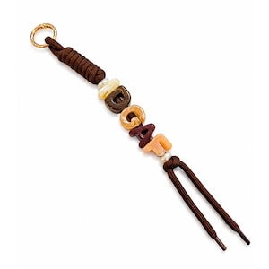 Peut inclure: Un porte-clés en cordon marron avec un anneau en métal doré en haut et des perles lettres formant le mot IDGAF dans des tons marron, beige et crème. Le cordon est noué autour des perles, avec deux extrémités libres dépassant en bas.