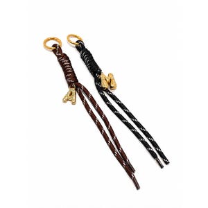 May include: Two decorative cord charms featuring gold-tone metal rings at the top. One cord is brown with white flecks and a gold-tone letter A charm, while the other is black with white flecks and a gold-tone letter M charm, both tied in knots.