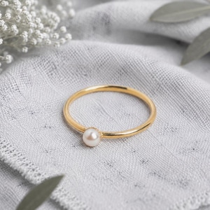 Op de afbeelding: Een dunne gouden ring met een enkele kleine, ronde, witte parel in het midden. De ring heeft een gladde, gepolijste afwerking en een eenvoudig, cirkelvormig ontwerp, wat het contrast tussen het gouden metaal en de witte parel benadrukt.
