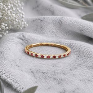 May include: A thin gold band ring featuring an alternating pattern of small, round, clear white stones and vibrant red stones set along the entire circumference of the metal band, creating a continuous, sparkling, and colorful circular design.