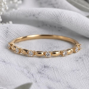 Op de afbeelding: Een dunne gouden ring met een herhalend patroon van kleine, gepolijste ovale gouden kralen afgewisseld met piepkleine, heldere, rondgeslepen edelstenen die langs de voorste helft van de metalen ring zijn gezet voor een verfijnd ontwerp.