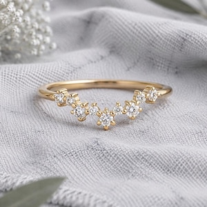 May include: A gold band ring featuring a curved, V-shaped design adorned with a series of small, clear, round-cut stones set along the top edge. The metal has a polished finish, and the stones are arranged in a cluster that follows the contour.