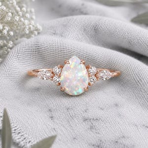 Op de afbeelding: Een roségouden ring met een centrale peervormige opaal. De opaal wordt geflankeerd door twee kleine ronde diamanten en één marquise-geslepen diamant aan elke kant, allemaal gezet in een delicate metalen band met een gepolijste afwerking.