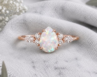 May include: A rose gold ring featuring a central pear-shaped opal gemstone. The opal is flanked by two small round diamonds and one marquise-cut diamond on each side, all set in a delicate metal band with a polished finish and prong settings.