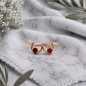 Könnte beinhalten: Ein goldfarbener Ring mit einer geschwungenen, abstrakten Silhouette, die zwei separate Schlaufen bildet, in denen jeweils ein kleiner, runder, tiefroter Edelstein sitzt. Die Ringschiene ist dünn und glatt und hat eine polierte Oberfläche.