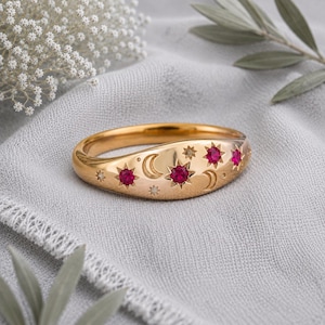 Op de afbeelding: Een goudkleurige ring met een hemels motief van gegraveerde maansikkels en sterpatronen. De ring is voorzien van vier kleine, ronde, felroze stenen die in het midden van de stervormige ontwerpen zijn geplaatst.