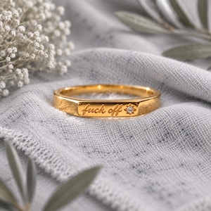 May include: A gold-toned metal ring featuring a flat, rectangular front surface engraved with the cursive text "fuck off" followed by a small, star-shaped accent containing a single, clear, round-cut gemstone set in the center of the star.