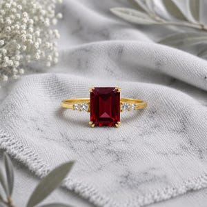 Peut inclure: Une bague en or ornée d'une grande pierre précieuse rouge foncé de taille émeraude rectangulaire, flanquée de deux petites pierres d'accent rondes et blanches transparentes de chaque côté, montée sur un anneau en or poli à quatre griffes.