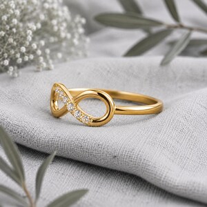 Op de afbeelding: Een gouden ring met een oneindigheidssymbool aan de voorzijde. Eén kant van de oneindigheidslus is ingelegd met vijf kleine, heldere, rondgeslepen stenen die het licht vangen, terwijl de rest van de ring een gladde, gepolijste gouden afwerking heeft.