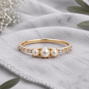 Könnte beinhalten: Ein Goldring mit einer zentralen Anordnung aus drei weißen Perlen, wobei die größte Perle in der Mitte von zwei kleineren flankiert wird. Der Ring ist mit kleinen, klaren, rund geschliffenen Steinen besetzt, die entlang der Goldschiene verlaufen.