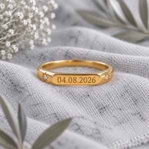Puede incluir: Un anillo de banda en tono dorado con una sección central plana y rectangular grabada con la fecha 04.08.2026. El anillo está decorado a ambos lados del panel central con un pequeño engaste en forma de estrella con una piedra clara.