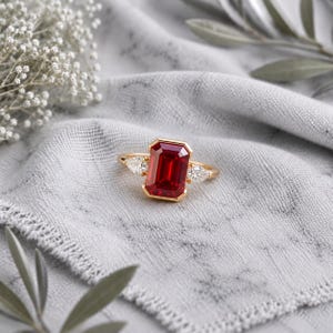 Puede incluir: Un anillo de oro con una gran piedra preciosa roja de corte esmeralda rectangular en el centro, flanqueada por dos piedras blancas transparentes más pequeñas en forma de pera a cada lado. El aro es fino y de oro pulido, con un engaste seguro.