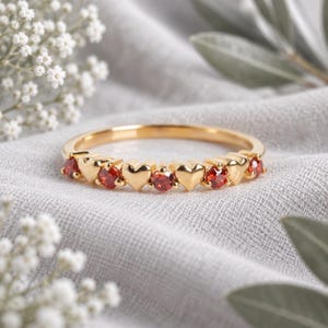 Op de afbeelding: Een gouden ring met een herhalend patroon van gepolijste gouden hartjes afgewisseld met kleine, ronde, gefacetteerde rode edelstenen. De ringband is dun en rond, met een verfijnd ontwerp en een heldere, reflecterende metalen afwerking.