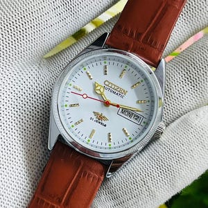 Op de afbeelding: Een Citizen automatisch polshorloge met een witte wijzerplaat, goudkleurige uurmarkeringen en een rode secondewijzer. Het horloge heeft een dag-datumvenster op de drie-uurpositie, een bruine lederen band met textuur en een zilverkleurige kast.