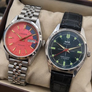May include: A Citizen automatic watch with a salmon-colored dial, silver-tone hands, and a metal link bracelet, alongside an HMT Pilot watch with a black dial, luminous markers, silver hands, and a black leather strap.