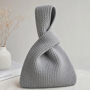 May include: A light grey crochet handbag featuring a triangular silhouette with a single integrated handle that loops through itself to create a secure closure. The bag is constructed from a dense, textured yarn with a consistent stitch pattern.