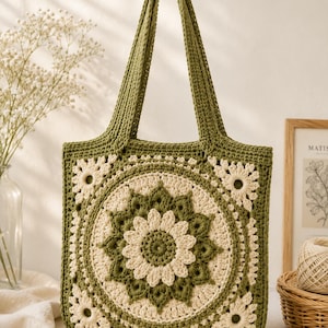 May include: A square crochet tote bag featuring a central circular floral motif in olive green and cream yarn. The bag has two matching olive green shoulder straps and a repeating flower pattern across the front panel, constructed with textured yarn.