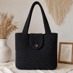 May include: A black crocheted tote bag featuring a rectangular body, two sturdy shoulder straps, and a central flap closure secured with a single brown wooden button. The bag is constructed with a dense, uniform stitch pattern throughout the exterior.