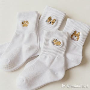 May include: A set of white crew socks featuring embroidered designs of a golden paw print, a corgi face, a corgi body, and a corgi rear end. The socks have a ribbed cuff and a plain white foot section, showcasing detailed stitching of the dog motifs.