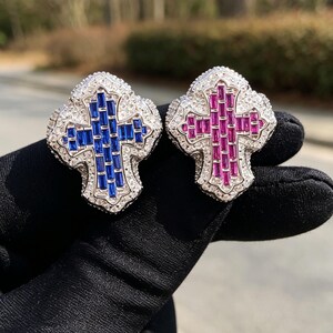 May include: Two cross-shaped pendants featuring silver-toned metal settings encrusted with small clear stones. One pendant displays a central inlay of rectangular blue stones, while the other features a central inlay of rectangular pink stones.