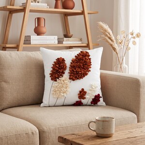 Peut inclure: Un coussin carré blanc orné d'un motif botanique décoratif composé de pétales en tissu superposés dans des tons orange rouille, crème et bordeaux profond. Les formes florales sont disposées en tiges verticales sur la face avant.