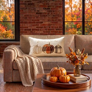 May include: A rectangular cream-colored throw pillow featuring the embroidered word pumpkin and three distinct pumpkin illustrations in orange, brown, and plaid patterns. The pillow rests on a sofa draped with a beige cable-knit throw blanket.
