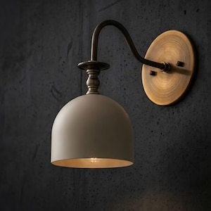 May include: A wall-mounted light fitting featuring a matte off-white dome-shaped shade suspended from a curved, antique brass-toned metal arm. The fitting includes a circular brass wall plate with a brushed finish and a decorative tiered neck.