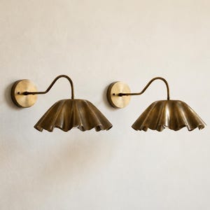 Op de afbeelding: Een paar wandlampen met een geborstelde messing afwerking. Elke lamp bestaat uit een ronde muurplaat, een gebogen arm en een brede, naar beneden gerichte kap met een opvallend golvend, geschulpt randontwerp in metallic messing.