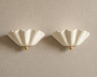 May include: A pair of wall sconces featuring off-white, ruffled, shell-like shades. Each fixture is accented with a small, polished gold-tone spherical detail at the base, providing a clean, symmetrical design for interior wall lighting applications.