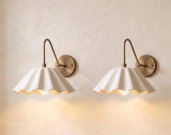 May include: A pair of wall sconces featuring white, scalloped, bell-shaped shades attached to curved, antique brass-toned metal arms. Each fixture includes a circular wall-mounting plate and emits a warm, soft glow from the base of the shade.
