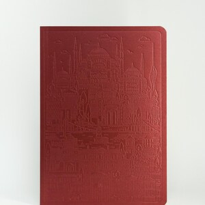 May include: A deep red notebook featuring an embossed line art illustration of a cityscape with multiple domed buildings, minarets, trees, and a bridge spanning across the lower section of the cover, with rounded corners on the right side.