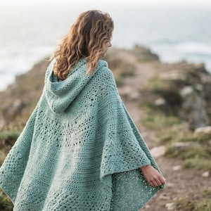 May include: A sage green hooded poncho featuring an intricate crochet pattern with a mix of solid stitches and openwork lace details. The garment has a loose, flowing silhouette with a pointed hemline and a hood attached at the neckline.