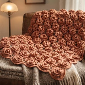 Puede incluir: Una manta de ganchillo color terracota con un patrón repetitivo de motivos de rosas tridimensionales. La manta tiene un borde festoneado y está colocada sobre un sillón de cuero marrón, sobre una manta tejida de textura en tonos neutros.