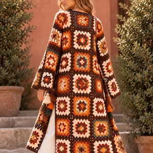May include: A long-sleeved duster cardigan made of brown-based granny square crochet patches in shades of orange, yellow, cream, and dark blue. The garment features a knee-length cut, wide sleeves, and decorative tassels hanging from the waist area.